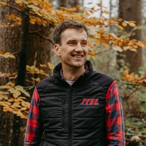 Man in the forest smiling, wearing a jacket with m3-ZT logo and striped shirt. Autumnal atmosphere.
