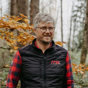 Man with glasses and striped shirt, surrounded by autumnal trees.